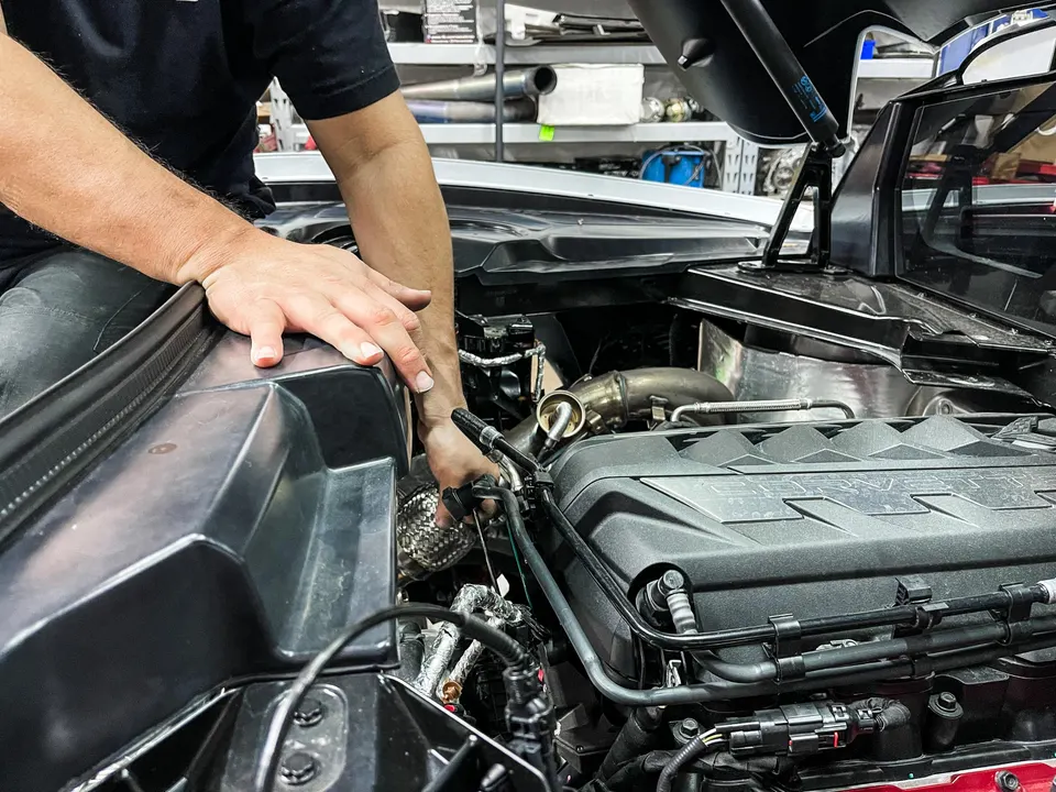 A mechanic's hands working on a Corvette C8 engine bay, installing a turbo system at a performance tuning shop.