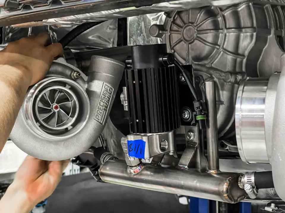 A close-up of a Precision Turbo turbocharger being installed in a car's engine bay, with hands visible during the process.