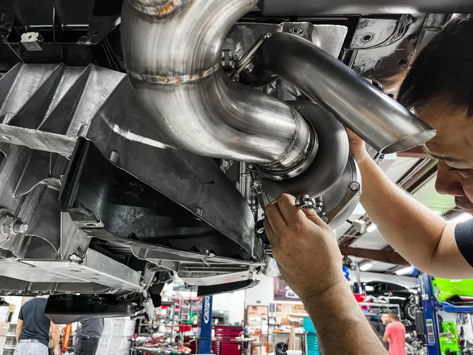 A mechanic installs a large turbocharger and custom boost piping on the underside of a performance car in a shop.