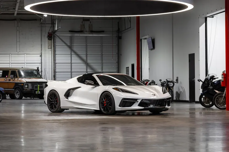 White Chevrolet Corvette C8 parked in a well-lit garage with other vehicles and motorcycles in the background.