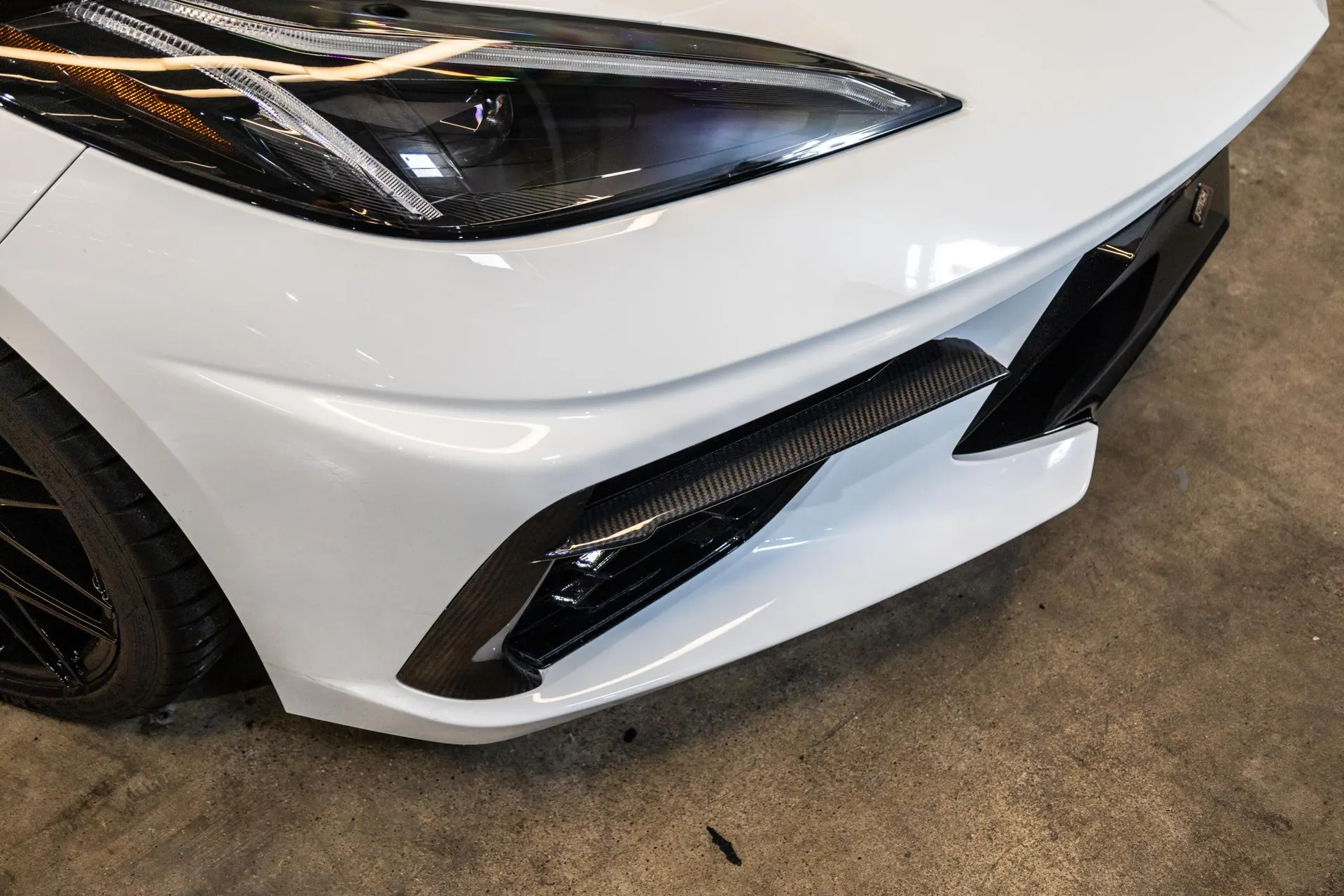 Close-up of a white Corvette C8 front bumper with carbon fiber aero accents and LED headlight.