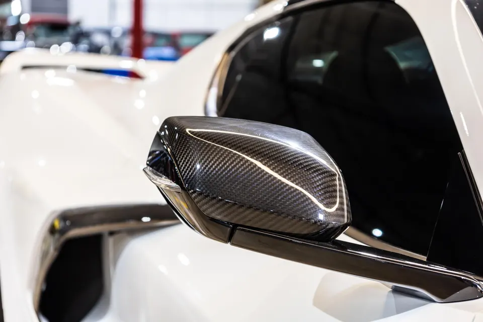 Close-up of a carbon fiber side mirror on a white luxury sports car, showcasing the weave pattern.