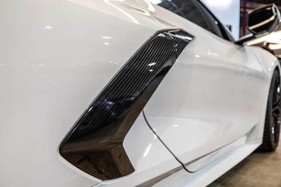 Close-up of a white Corvette's carbon fiber side fender vent, showcasing the weave pattern and glossy finish.