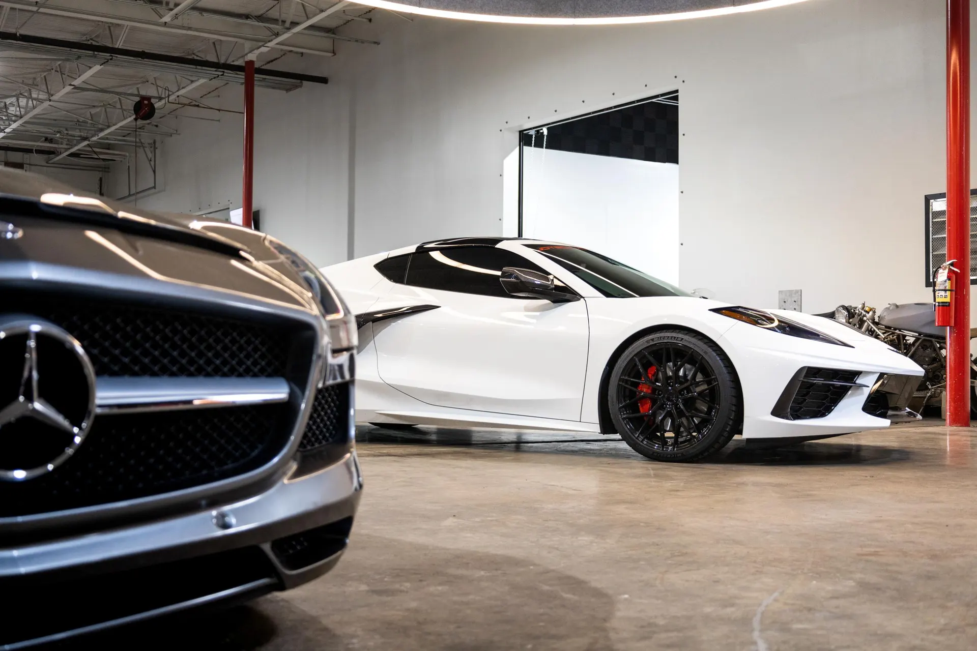 A white Corvette C8 and a Mercedes-Benz parked inside a clean automotive shop or garage with concrete floors.
