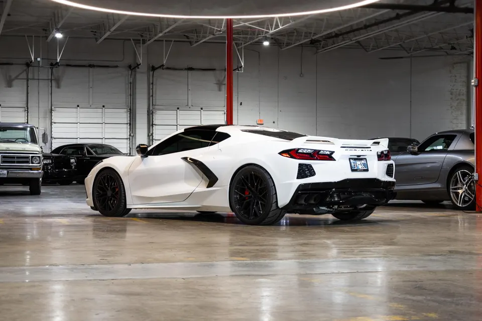 A white Chevrolet Corvette C8 with black wheels parked in a clean garage with other vehicles in the background.