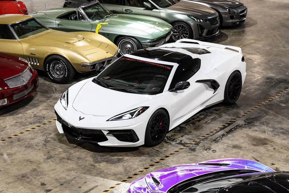 Overhead view of a white C8 Corvette surrounded by classic Corvettes and other luxury cars in a Jotech Motorsports showroom.