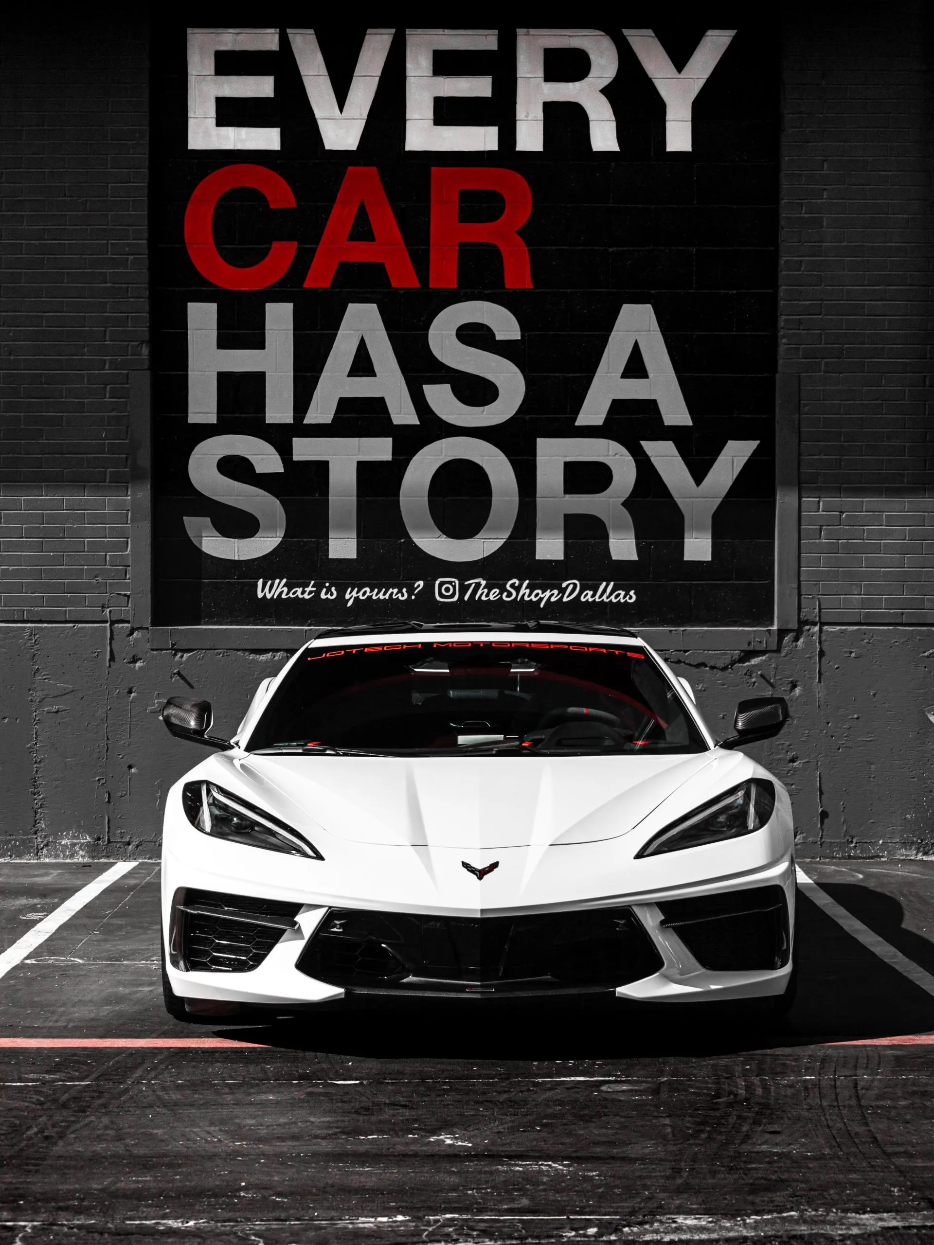 A white Corvette C8 parked in front of a black brick wall with "EVERY CAR HAS A STORY" painted on it.