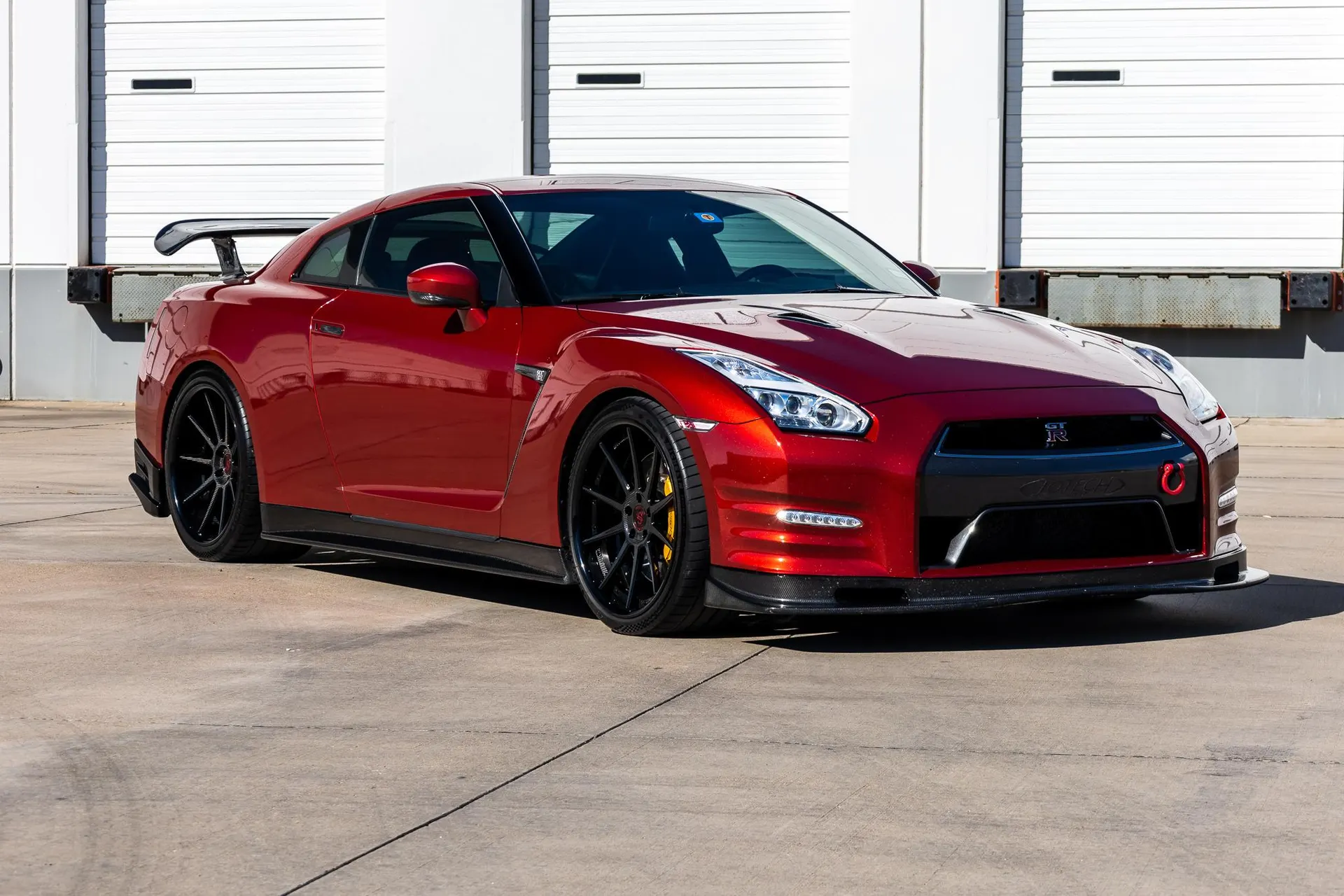 A red Nissan GT-R sports car with black aftermarket wheels and a carbon fiber rear spoiler parked in front of white garage doors.