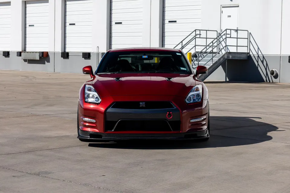 A red Nissan GT-R sports car parked in front of a warehouse with loading docks on a sunny day, showcasing its front profile.