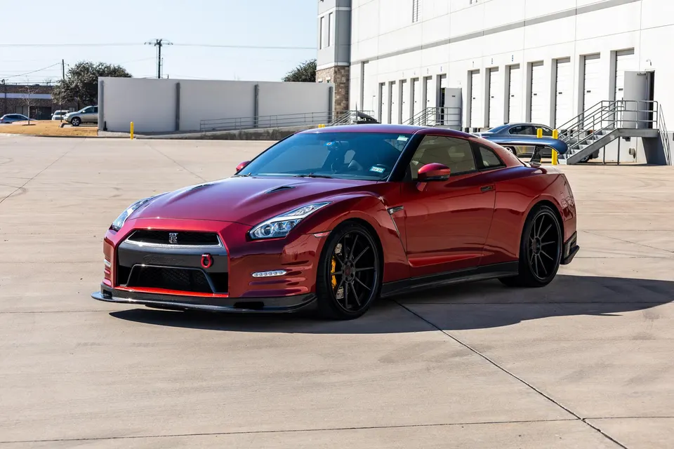A red Nissan GT-R with black aftermarket wheels and a carbon fiber front splitter parked in a sunny industrial lot.