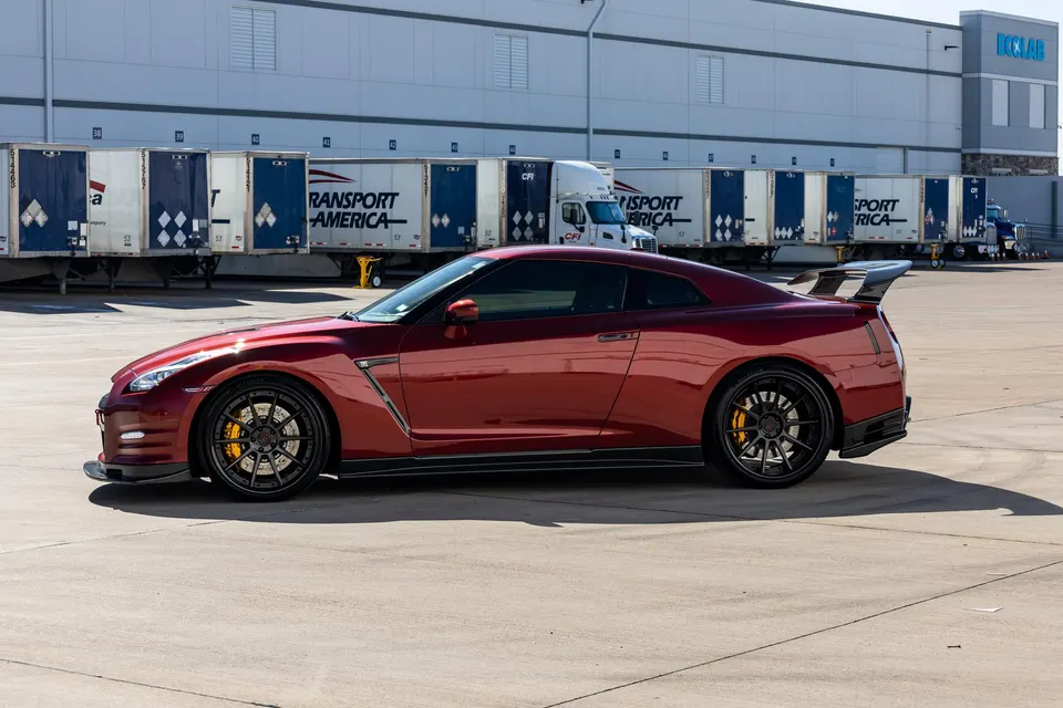 A red Nissan GT-R with aftermarket wheels and a large rear wing parked in front of a warehouse with semi-trailers.