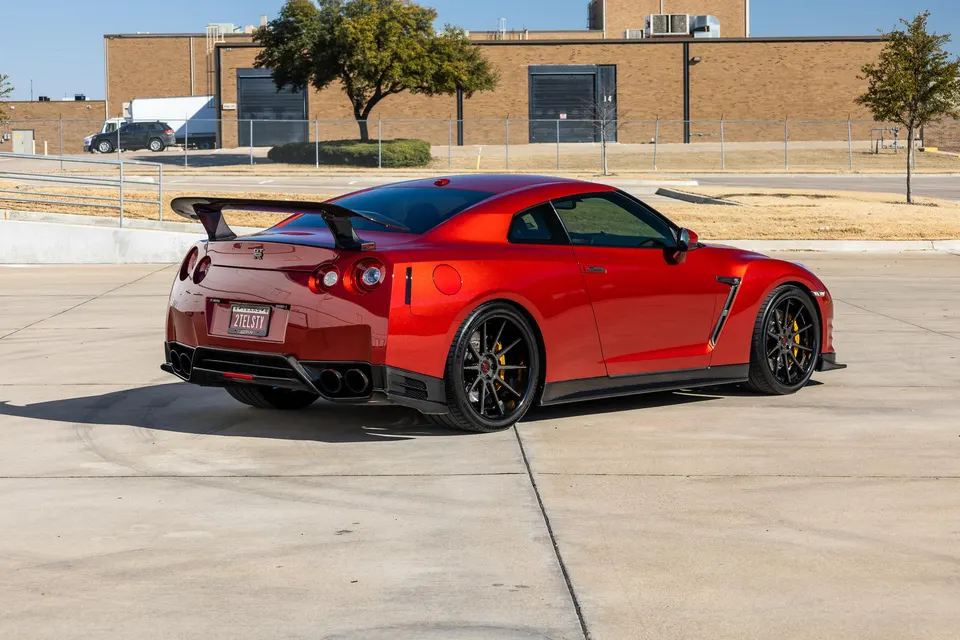 A red Nissan GT-R with a large carbon fiber wing and aftermarket black wheels parked outdoors on a sunny day.
