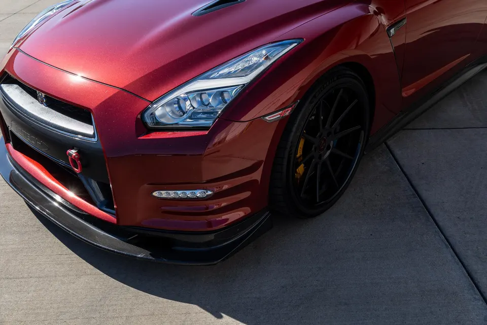 A close-up of a red Nissan GT-R's front corner, featuring a carbon fiber front splitter, black aftermarket wheels, and a red tow hook.