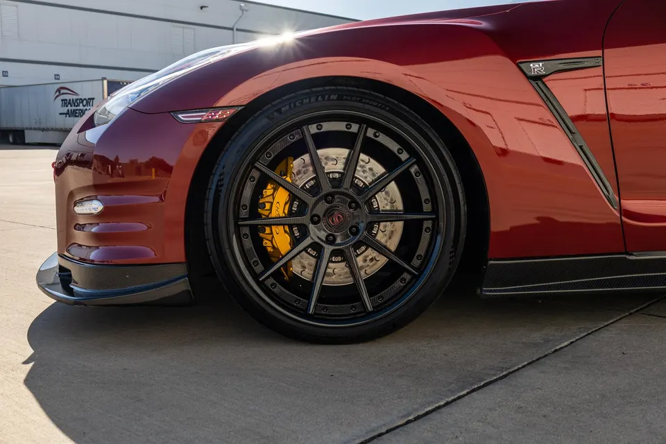 A close-up detail shot of a red Nissan GT-R's custom black wheel with yellow brake calipers and carbon fiber aero.