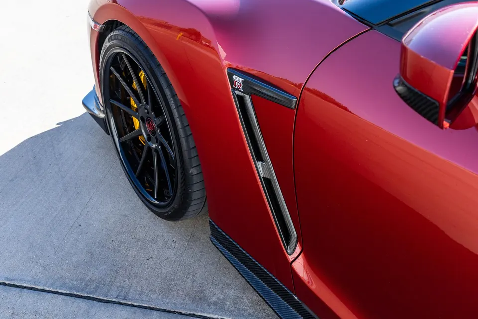 Close-up of a red Nissan GT-R's front wheel, carbon fiber fender vent, and side skirt with yellow brake calipers.