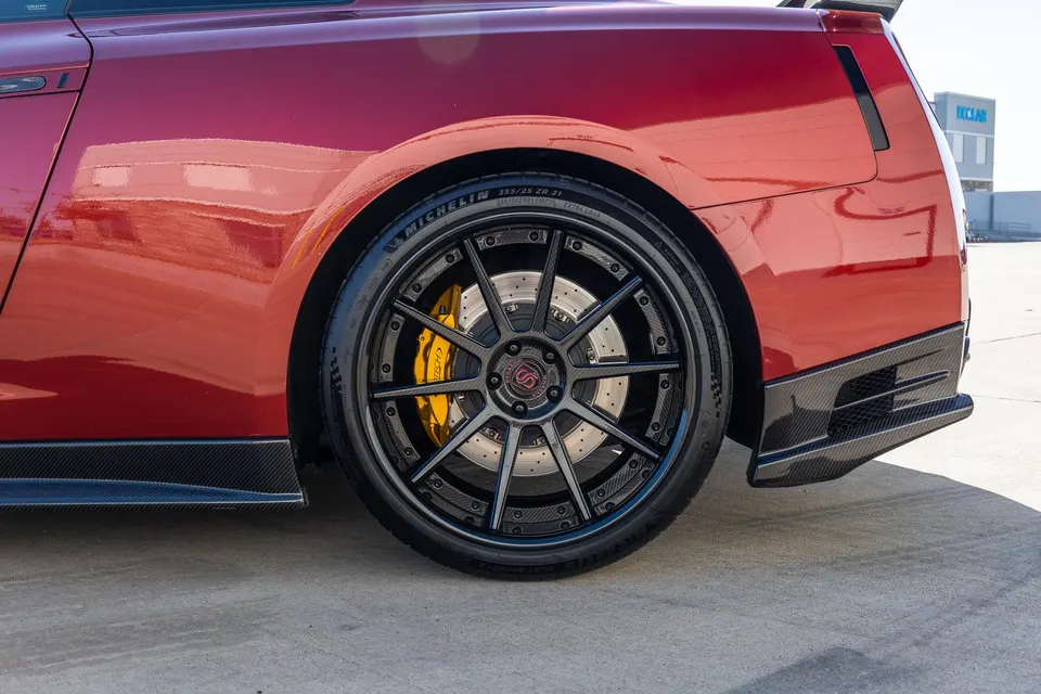 A close-up of a red Nissan GT-R's rear wheel, featuring black custom wheels, yellow brake calipers, and carbon fiber aero.
