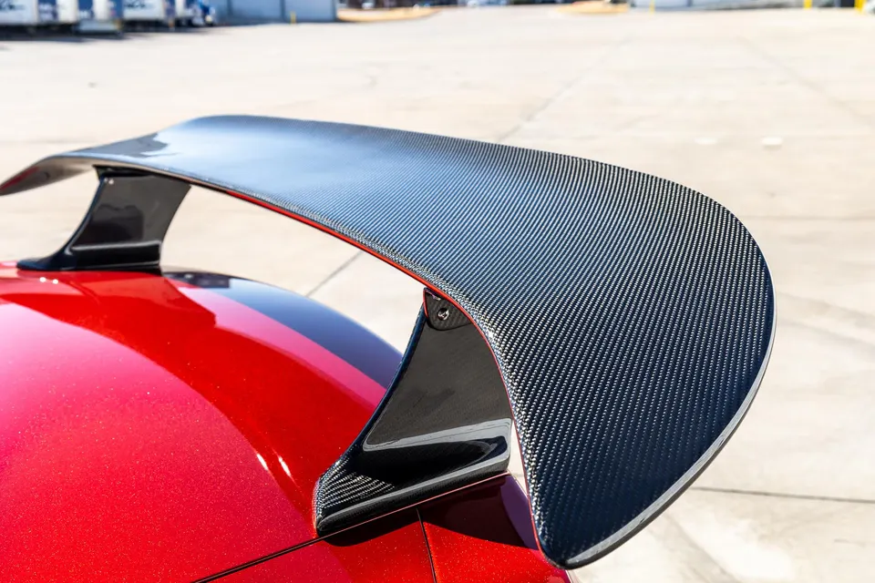 Close-up of a carbon fiber rear wing on a red sports car, highlighting the weave pattern and aerodynamic design.