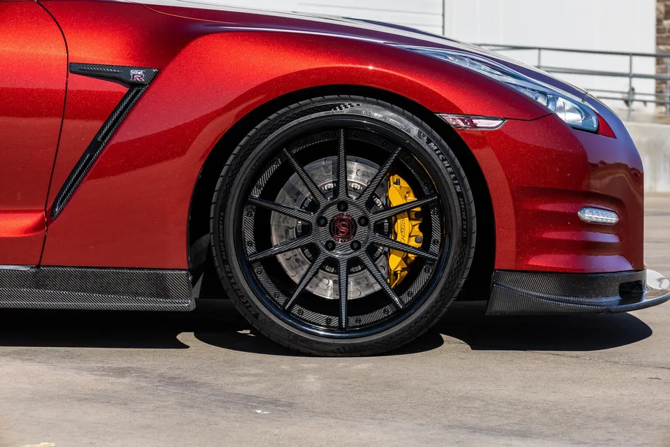 Close-up of a red Nissan GT-R's front wheel, featuring custom black carbon fiber wheels, yellow brake calipers, and carbon fiber aero.