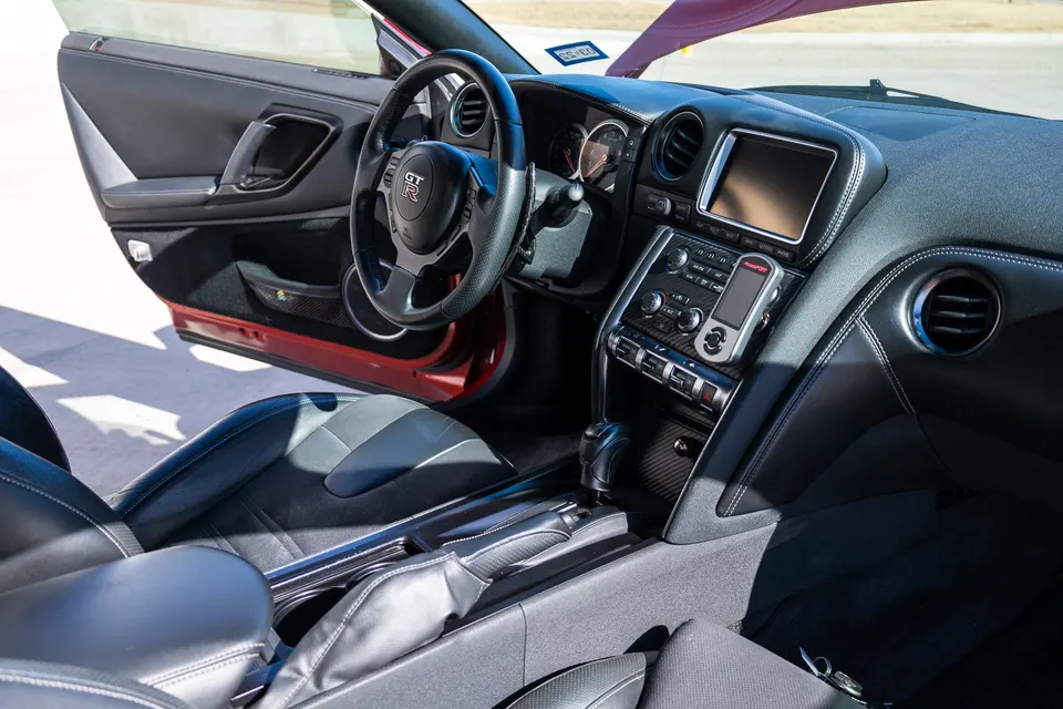 Interior view of a Nissan GT-R showing the dashboard, steering wheel, center console, and a mounted Cobb Accessport.