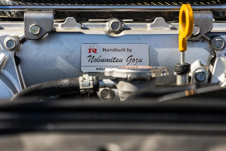 A close-up of a Nissan GT-R engine bay showing the 'Handbuilt by Nobumitsu Gozu' plaque and yellow dipstick.