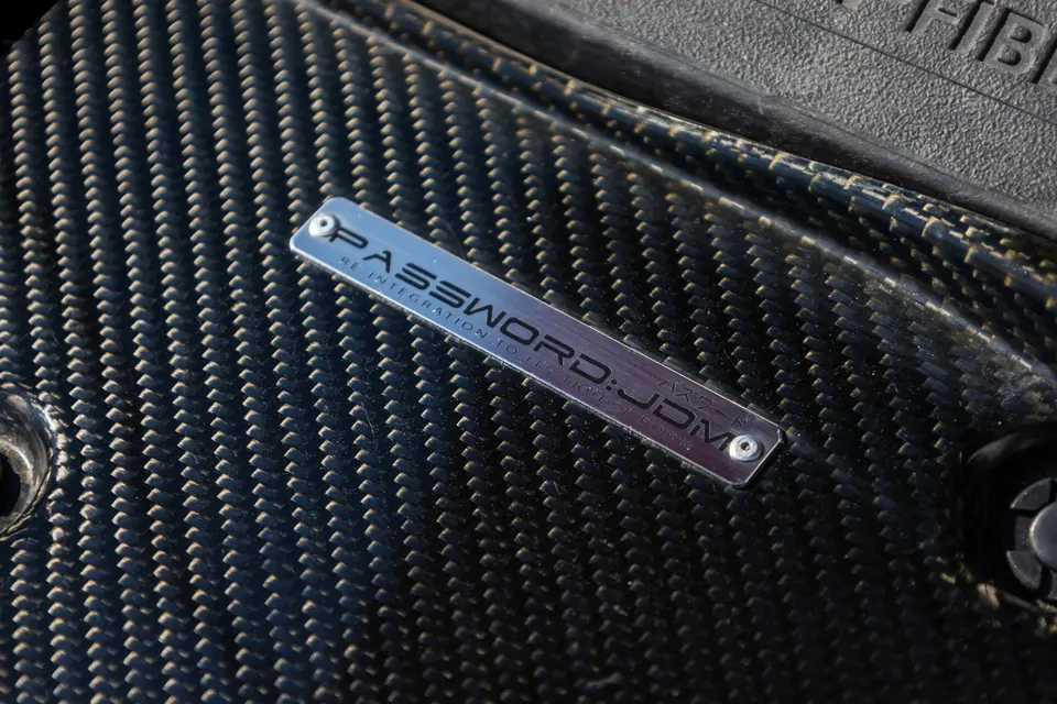 Close-up of a Password:JDM carbon fiber part with a metallic badge, showcasing the weave and branding.