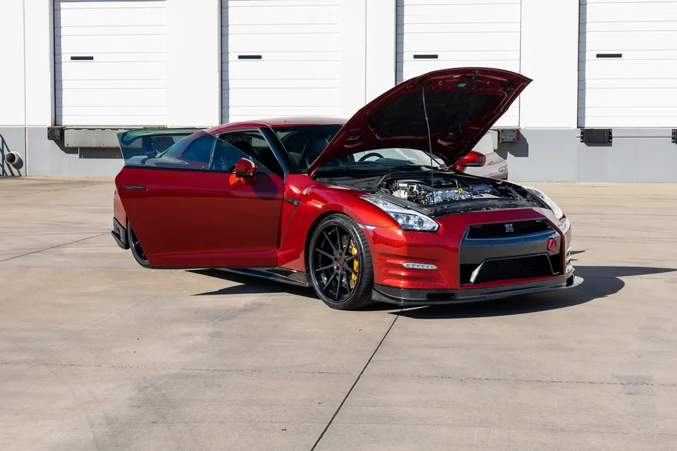Red Nissan GT-R R35 with hood and driver's door open, showcasing the engine bay and custom wheels in front of a shop.