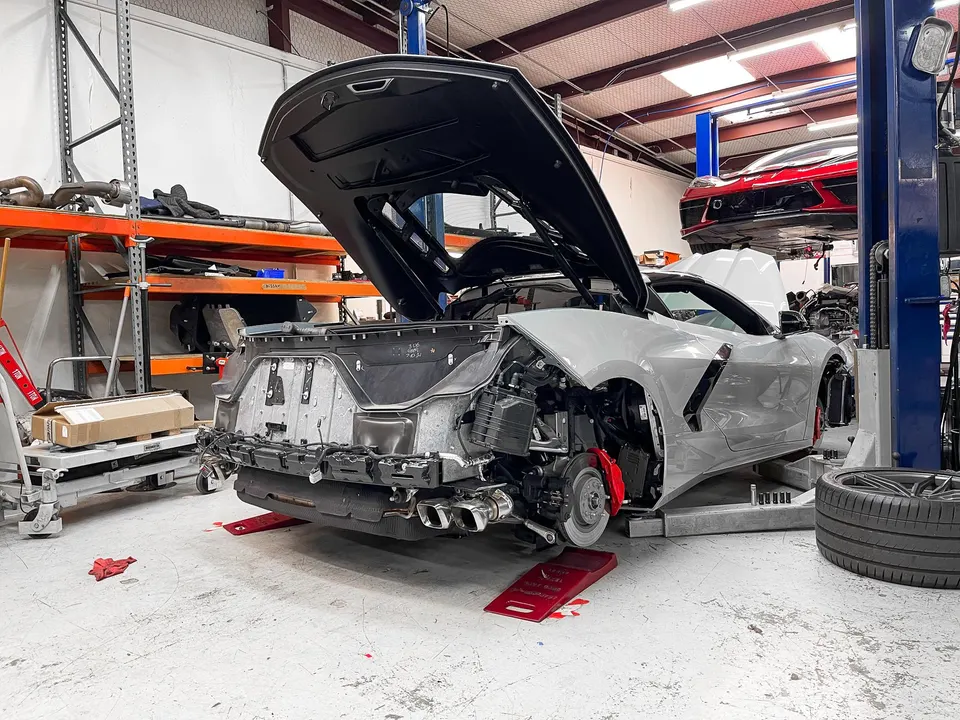 A grey Corvette C8 undergoing disassembly in a performance tuning shop with its engine bay open and rear bumper removed.