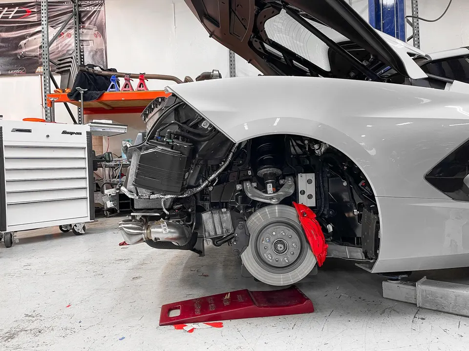 White C8 Corvette with front fender removed, showcasing red brake calipers and suspension components in a performance shop.
