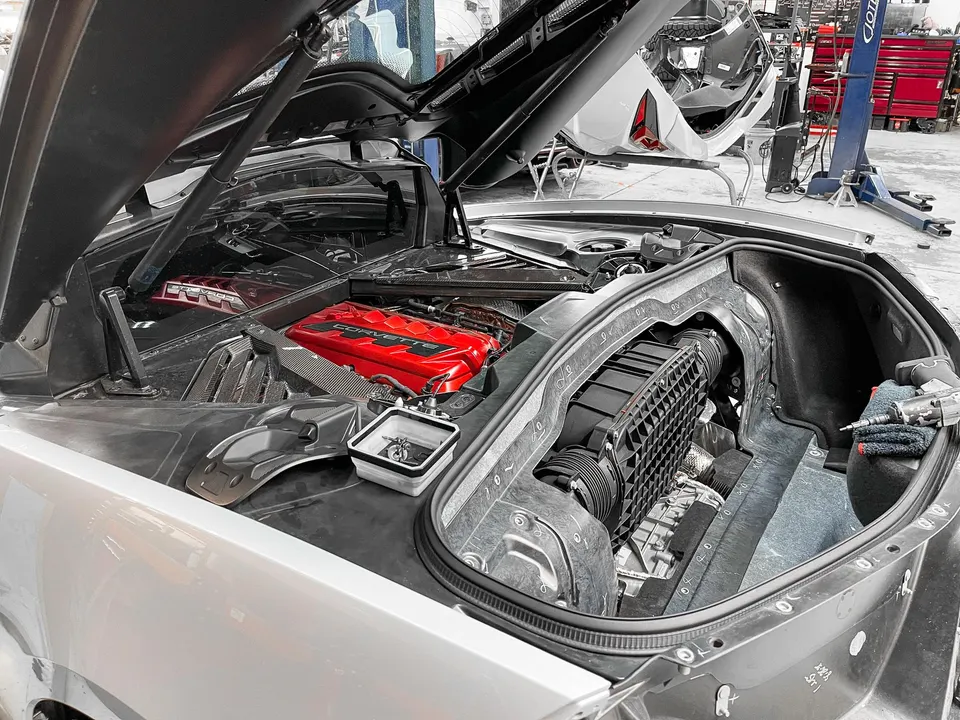 A silver Corvette C8 with its engine bay open in a shop, showcasing the red engine cover and air intake system.
