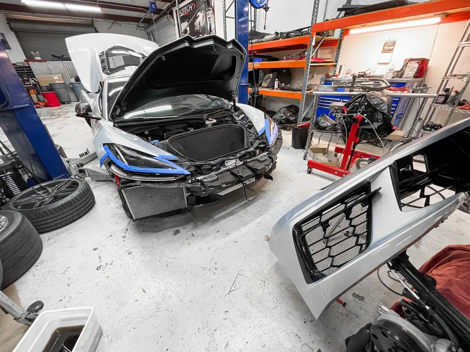 A silver Corvette C8 undergoing work in a performance shop with its front bumper removed, revealing the radiator.