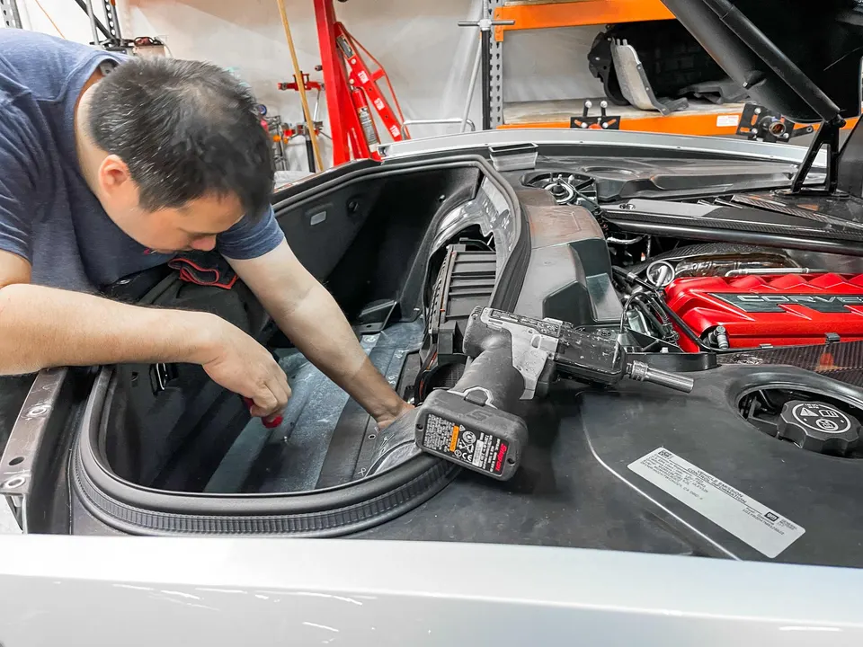 Mechanic working on the engine bay of a silver Corvette C8 in a performance tuning shop.