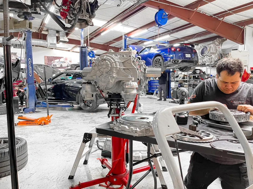 A mechanic works on a drivetrain component in a busy performance tuning shop with multiple Nissan GT-Rs on lifts.