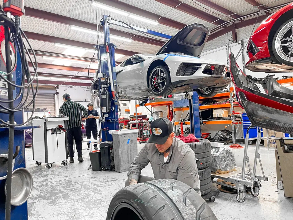 Mechanics working on a white Corvette C8 on a lift and other performance cars in a busy Jotech Motorsports shop.