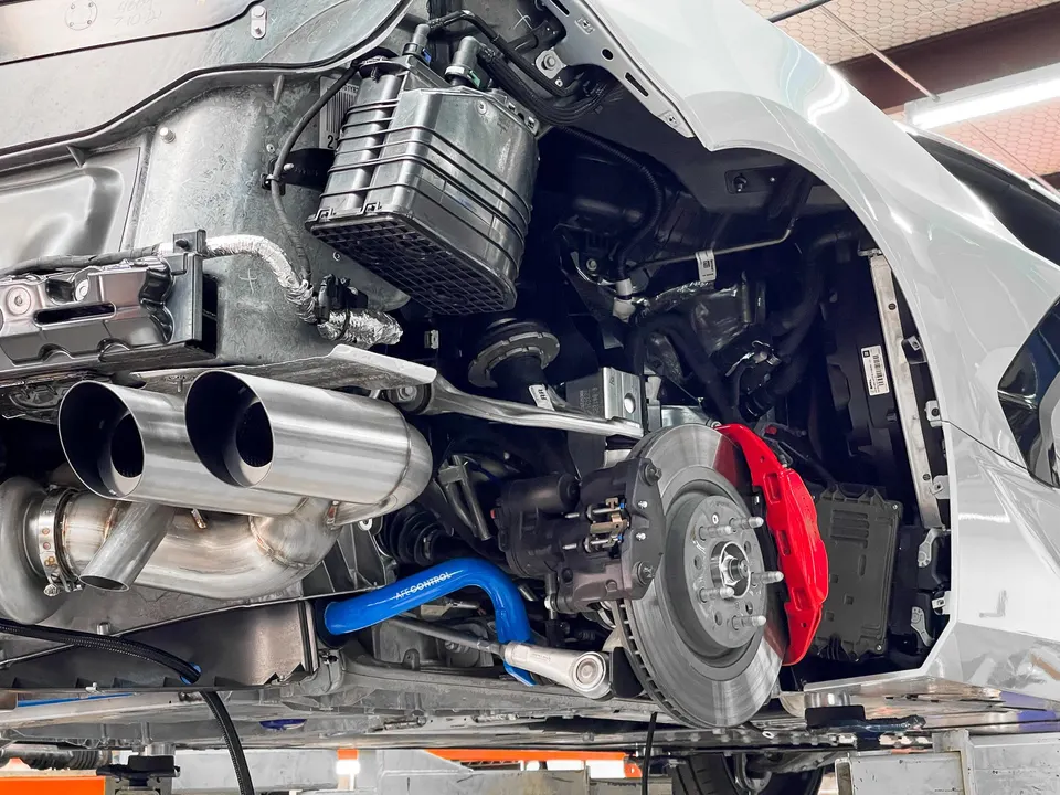 Underside view of a white Corvette C8 on a lift, showcasing an aftermarket exhaust system, red brake calipers, and an AFE Control sway bar.