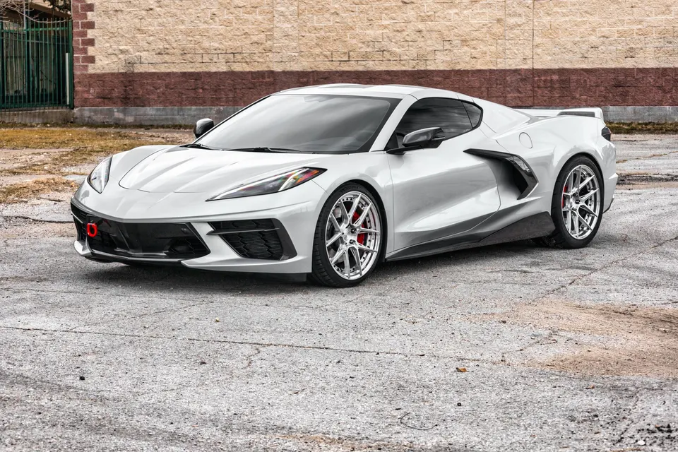 White Chevrolet Corvette C8 with aftermarket chrome wheels, red brake calipers, and a red tow hook parked outdoors.
