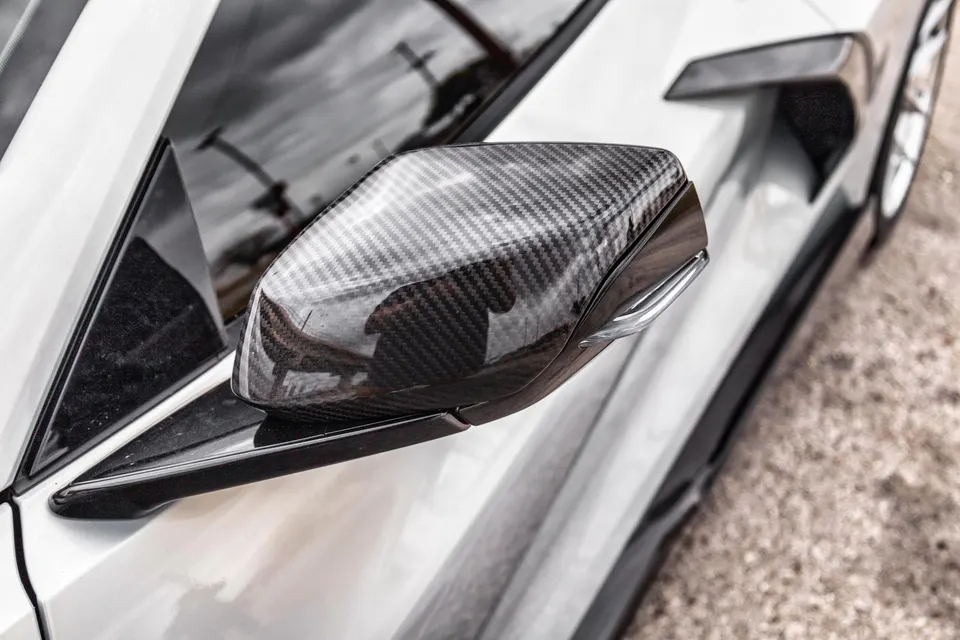 Close-up of a white car's carbon fiber side mirror, showcasing the weave pattern and sleek design.