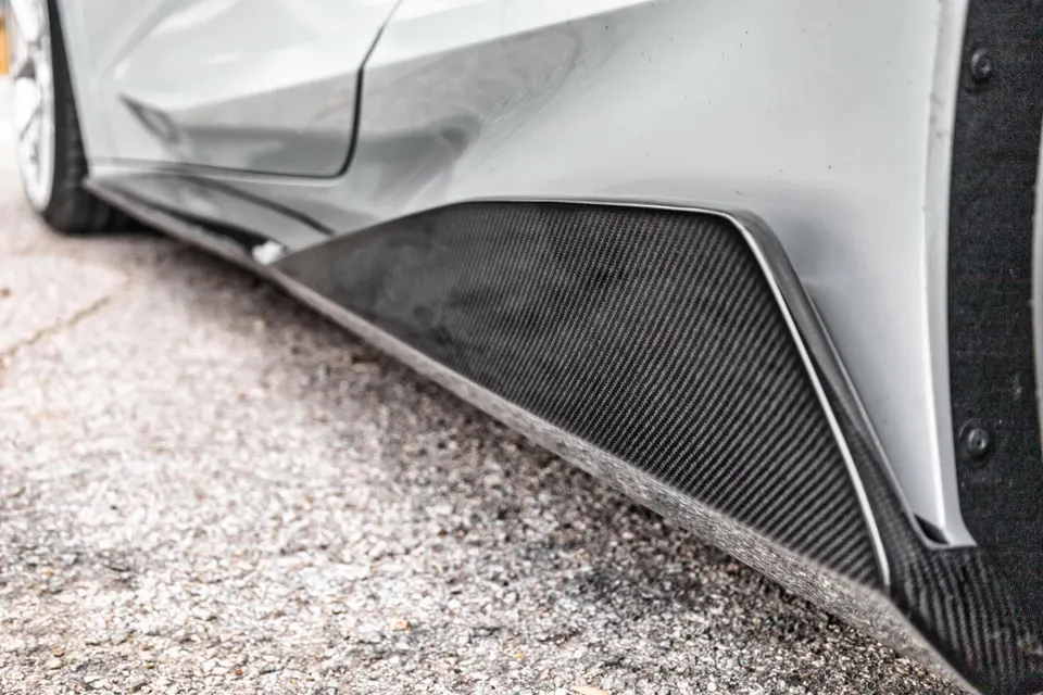 Close-up of a carbon fiber side skirt on a grey performance car, highlighting the weave pattern and aerodynamic design.