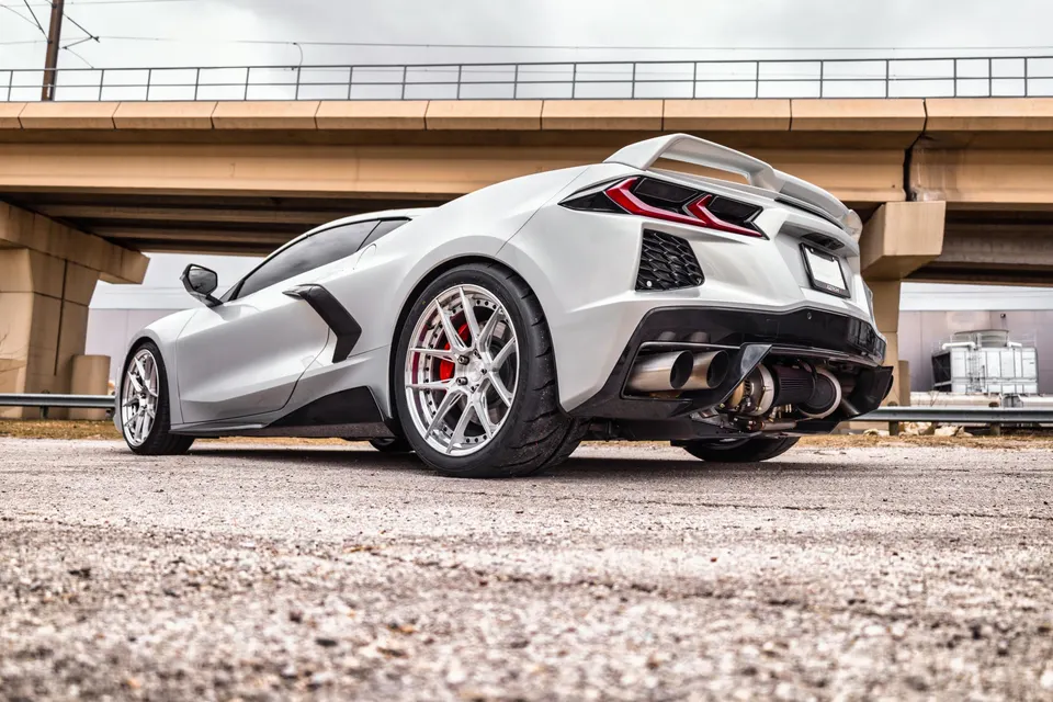 A white Corvette C8 with aftermarket wheels and a custom exhaust system featuring a visible turbocharger, parked under an overpass.