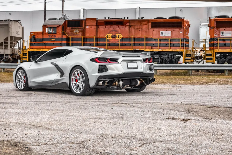 Silver Corvette C8.R with aftermarket exhaust and custom wheels parked in front of an orange train.