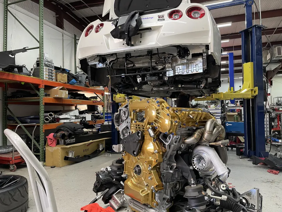 A white Nissan GT-R on a lift in a performance shop, with its gold-painted engine and large turbocharger removed and on a stand.
