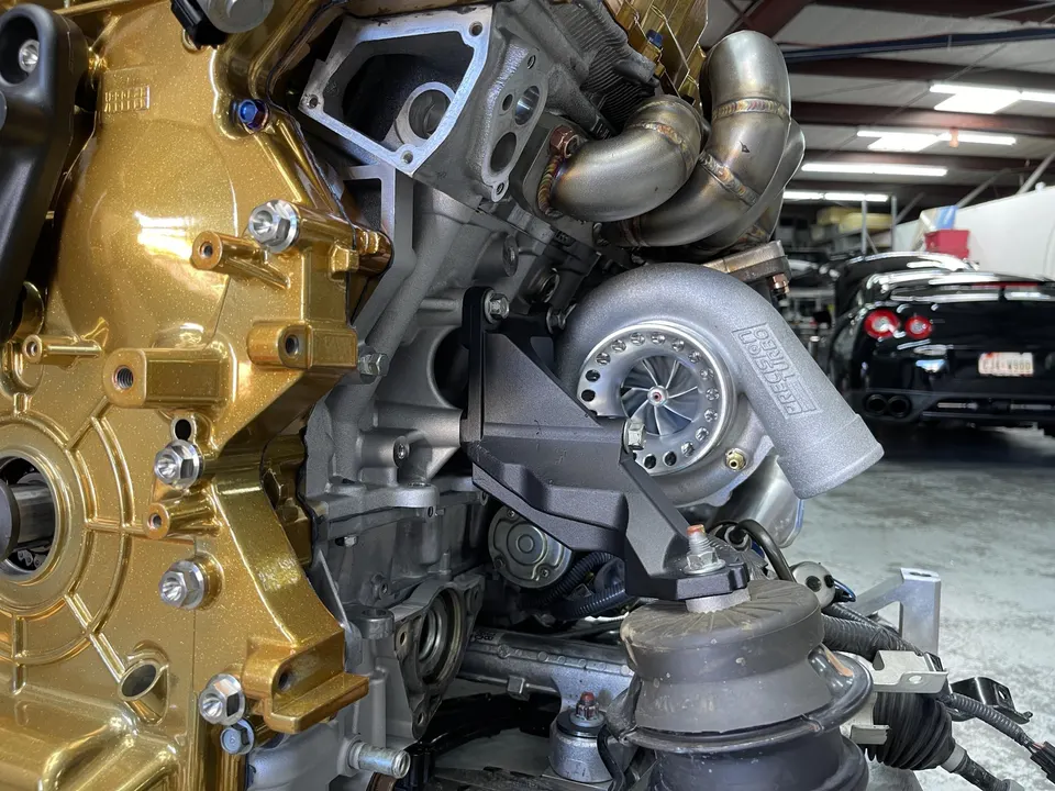 Close-up of a gold-painted engine block with a Precision Turbo turbocharger and custom exhaust manifold in a shop.