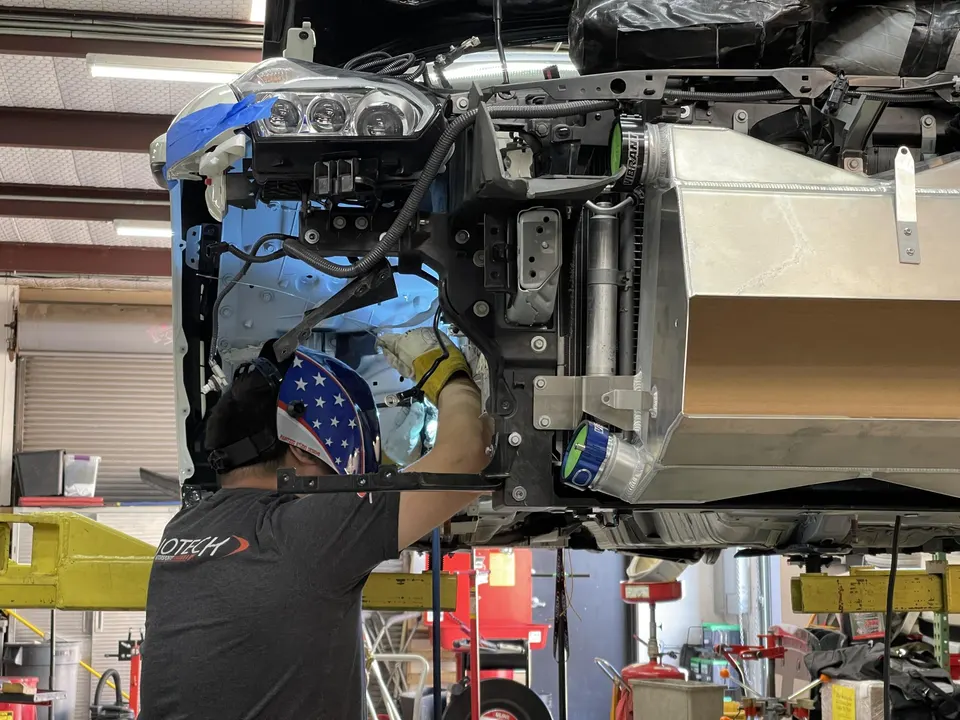 A mechanic in a stars and stripes welding helmet works on the front end of a car on a lift, installing a large aftermarket intercooler.