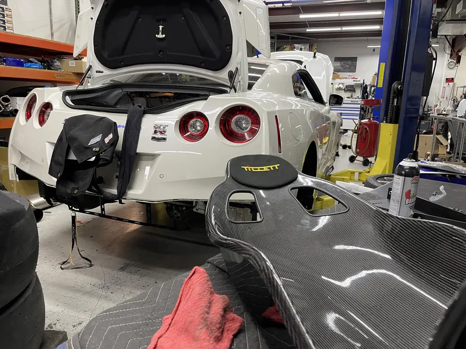 White Nissan GT-R on a lift in a performance shop with a carbon fiber racing seat in the foreground.