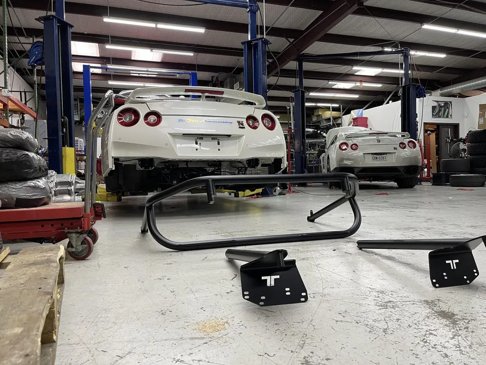 Two white Nissan GT-R R35s in a performance tuning shop with a rear chassis brace on the floor.