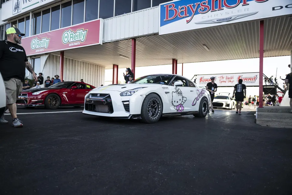 A white Nissan GT-R with a Hello Kitty wrap and a red Nissan GT-R in the staging lanes at a drag strip event.