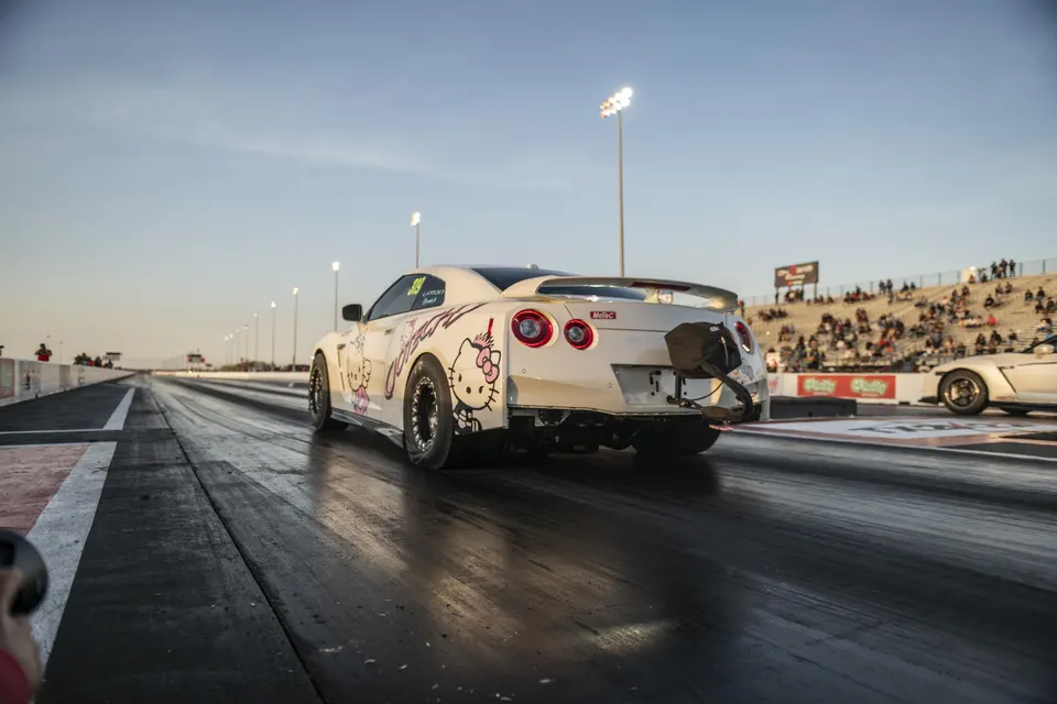 White Nissan GT-R with Hello Kitty decals on a drag strip, featuring drag slicks and a parachute at the rear.