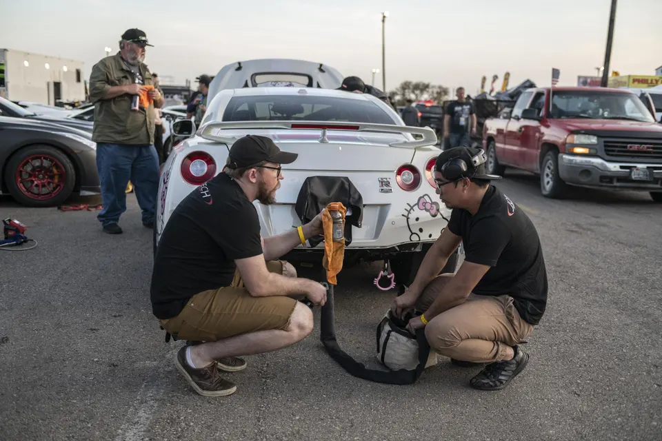 Two men preparing a white Nissan GT-R with Hello Kitty decals at a track event, with other cars in the background.