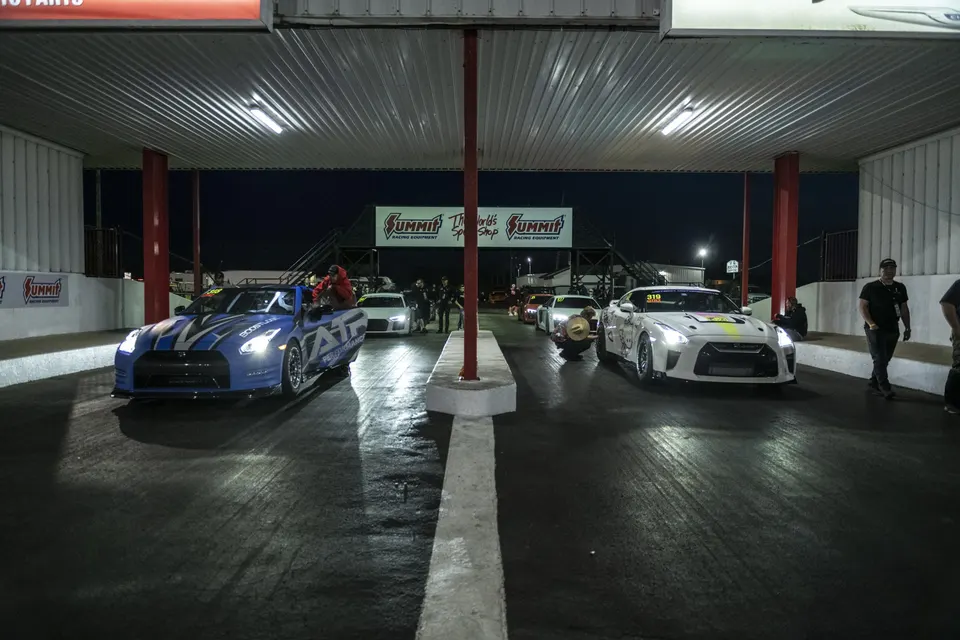Two Nissan GT-Rs, one blue and one white, in the staging lanes at a drag strip at night, ready to race.
