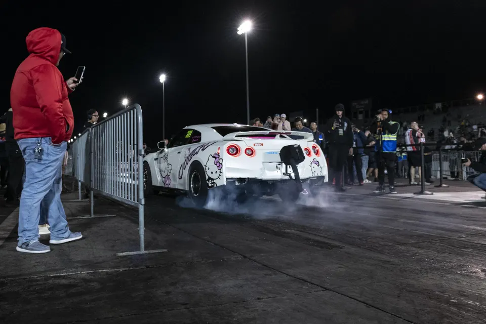 White Nissan GT-R with Hello Kitty livery performing a burnout on a drag strip at night with spectators.