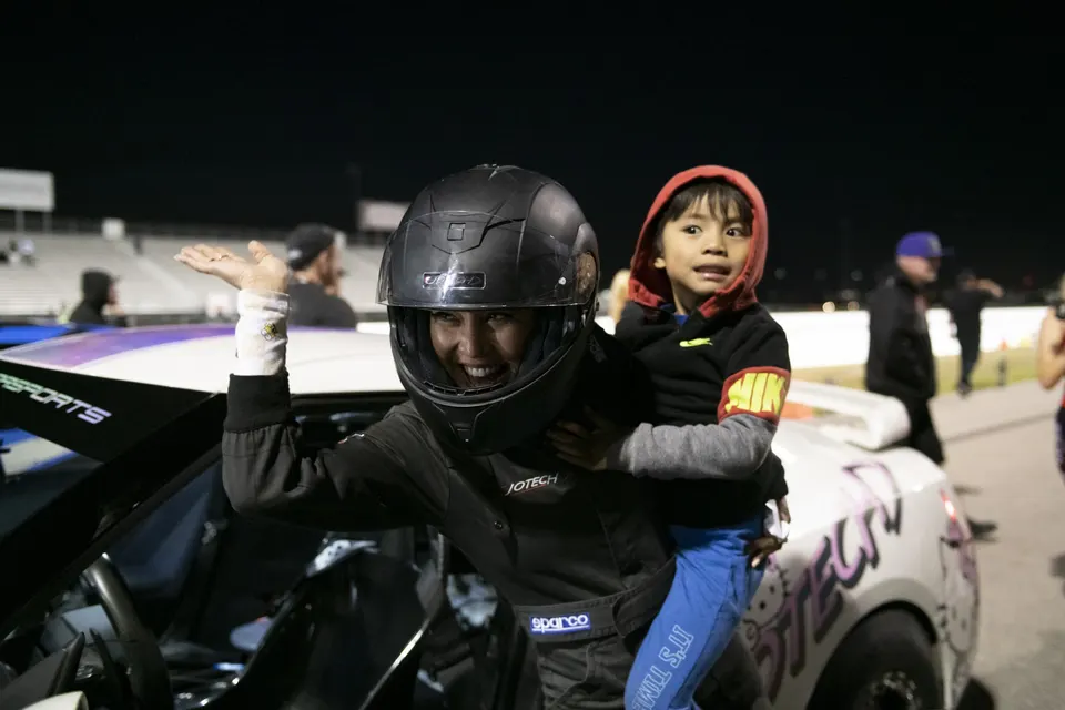 A smiling racer in a helmet and suit with a child on her back at a night drag strip event.